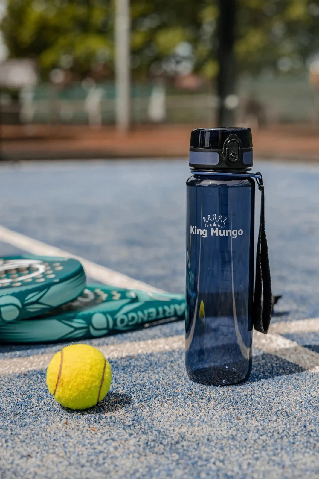 1 liter drinkfles van King Mungo op padelbaan, naast padel rackets en tennisballen.